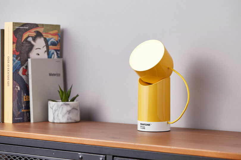 A yellow light labeled "PANTONE 7408 C" sits on a wooden shelf beside books and a small plant in a marble pot against a neutral wall.