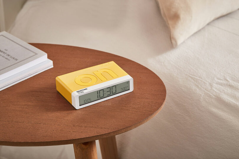 A digital alarm clock with a yellow casing sits on a wooden bedside table next to some books and a bed.