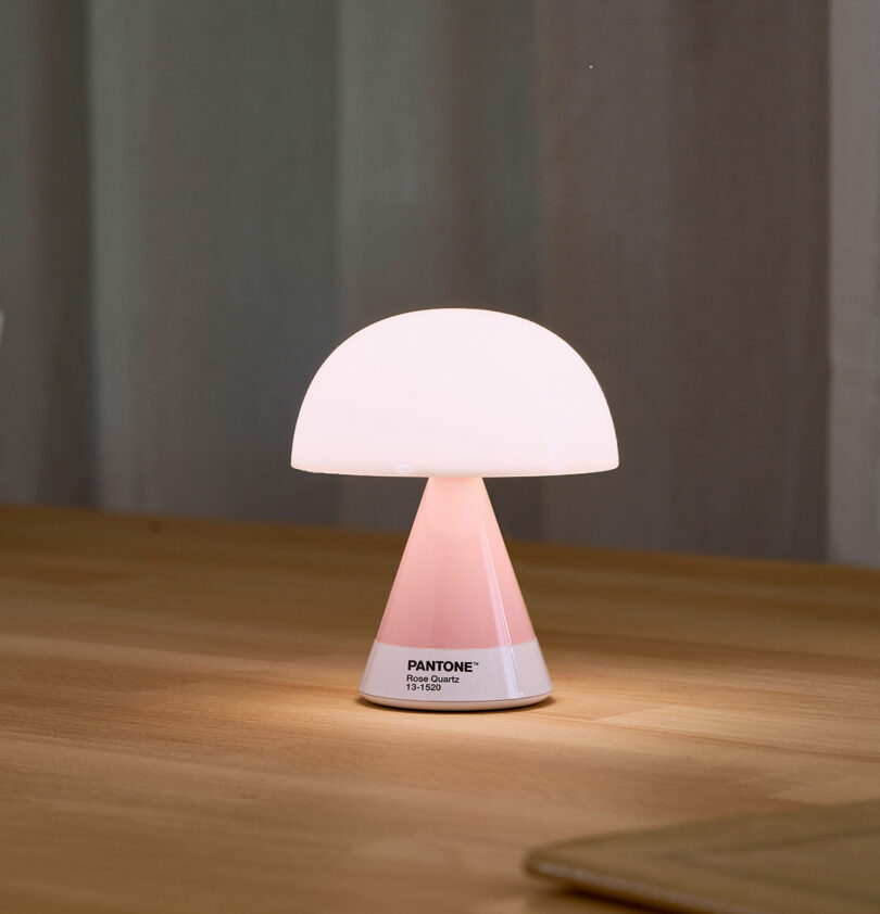 A small, mushroom-shaped table lamp with a pink base and white dome shade, labeled "Pantone 13-1520 TPG" on its base, sits on a wooden surface.