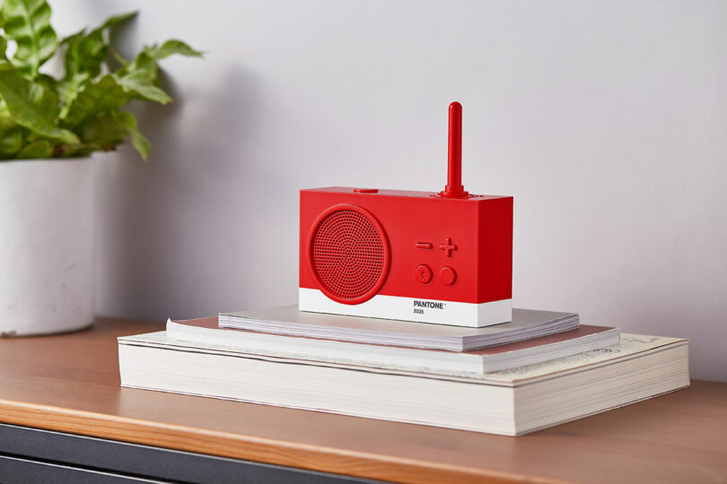 A red and white Pantone radio with an antenna sits on a pile of books on a wooden surface next to a potted plant.