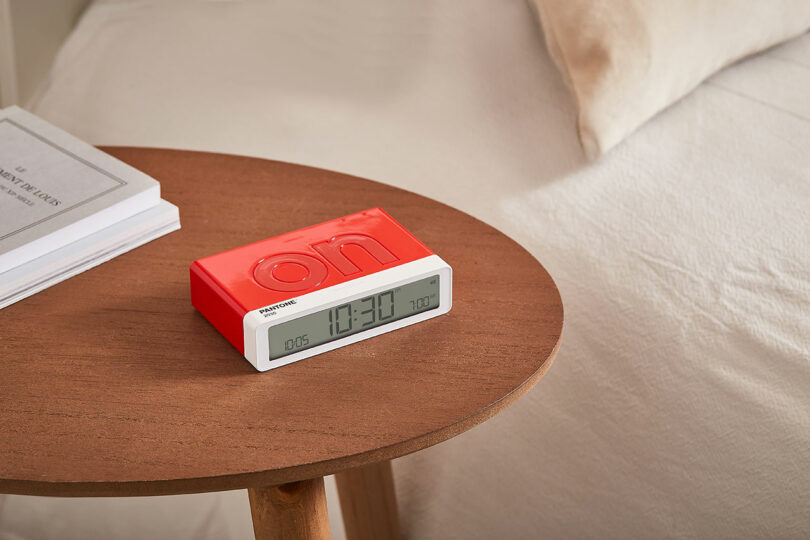 A red digital alarm clock displaying the time 10:30 AM sits on a round wooden bedside table next to a stack of books.