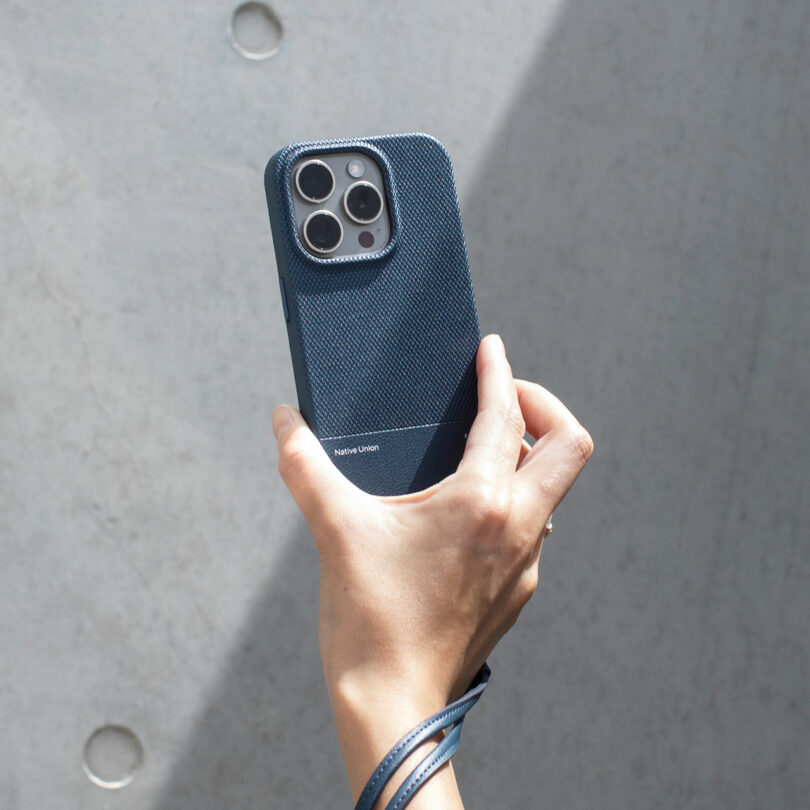 A hand holds a smartphone in a textured dark blue case with minimal branding by Native Union, against a plain gray background.