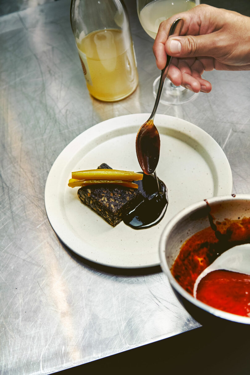 A hand uses a spoon to drizzle dark sauce onto a dish containing grilled food and yellow vegetables on a white plate. A bottle and glass with yellow liquid are in the background on the stainless steel surface.