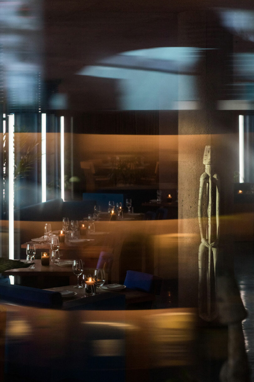 A dimly lit modern restaurant interior with set tables, chairs, and a lit candle on each table at Desa Potato Head. A reflective surface creates light streaks and a blurred figure is visible in the background.