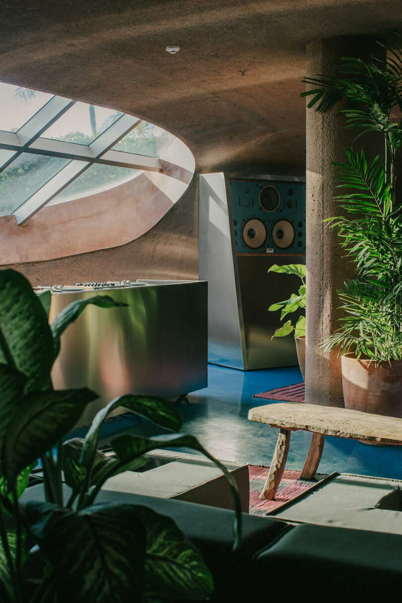 A modern interior with large windows, a futuristic DJ booth, lush green plants, and a wooden bench. Sunlight streams in, creating a warm and inviting ambiance.