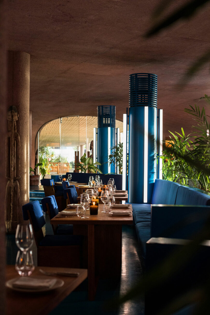 A dimly lit restaurant features blue padded seating with wooden tables set with wine glasses, plates, cutlery, and candles, surrounded by plants and modern blue columns.