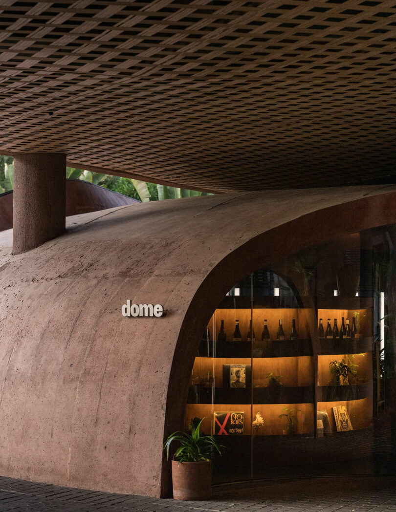 A curved concrete structure with a lattice roof stands outside the building. A sign reading "dome" is visible on the wall, and through the glass windows of Desa Potato Head, shelves filled with various bottles and decor can be seen.