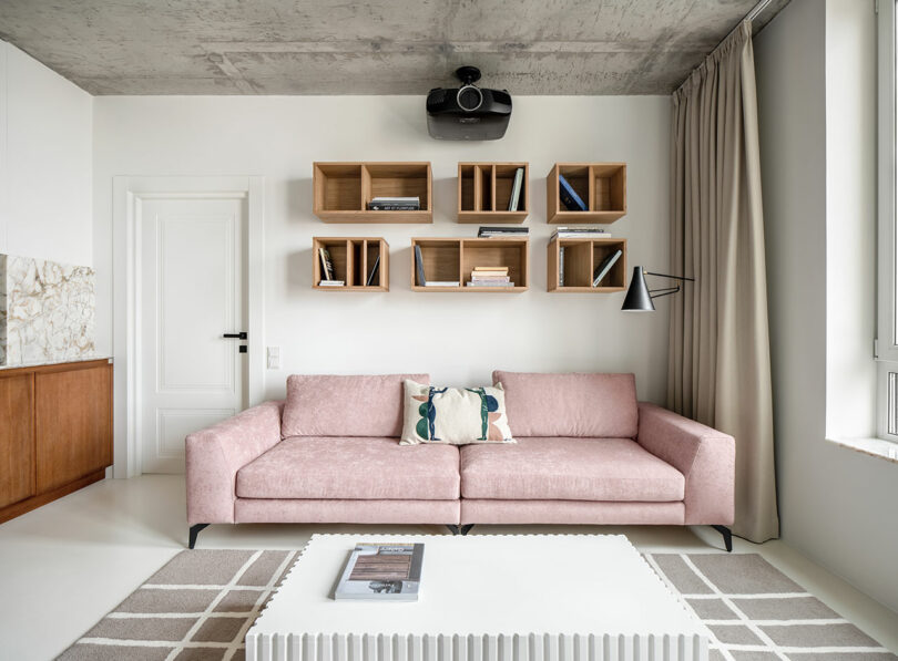 Modern living room featuring a pink sofa, white coffee table, and a wall-mounted shelving unit with books and decor items. A ceiling-mounted projector and floor-to-ceiling curtains are also visible.