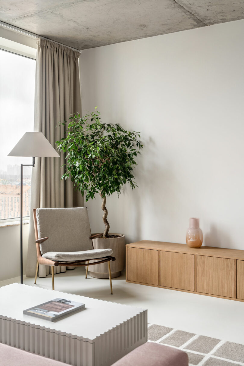 A minimalist living room with a beige armchair, a floor lamp, a potted plant, a wooden cabinet, and a white coffee table holding a magazine.