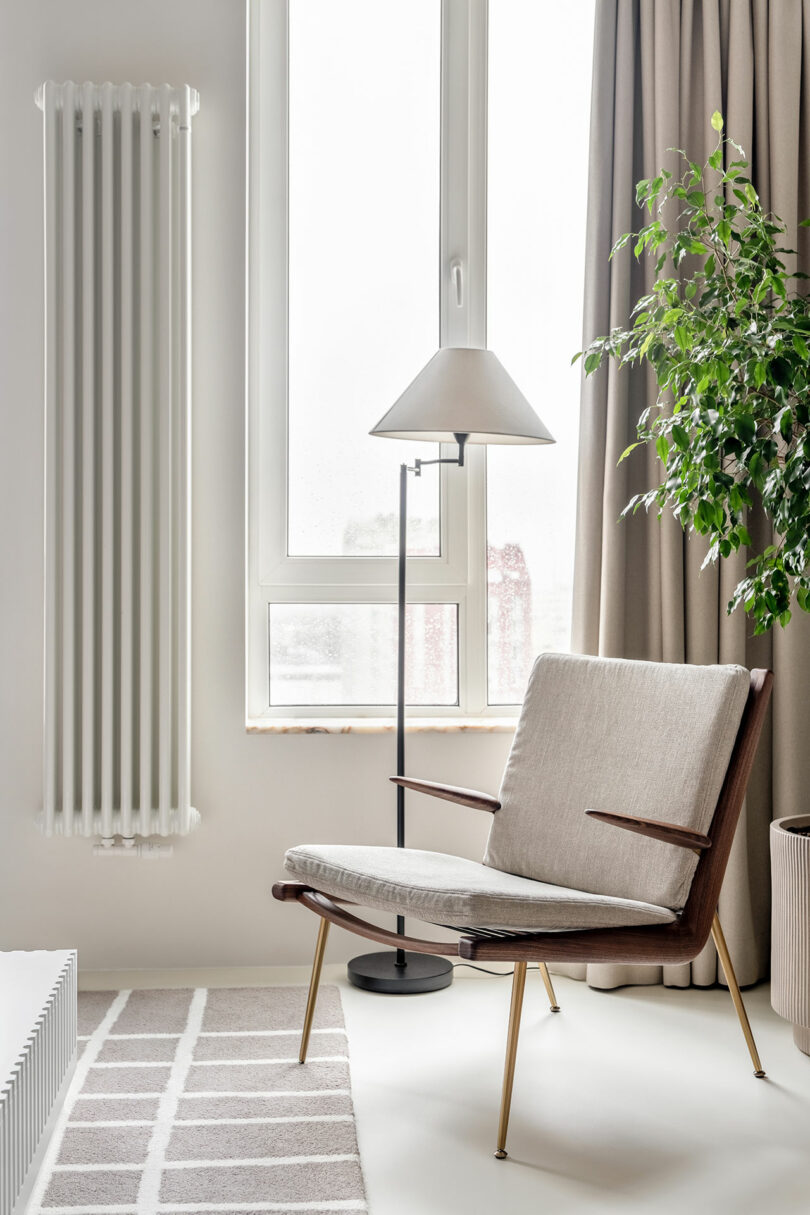 A minimalist room featuring a beige armchair, a standing lamp, a tall radiator, and a potted green plant beside a large window with beige curtains. The floor has a light-colored rug with geometric patterns.