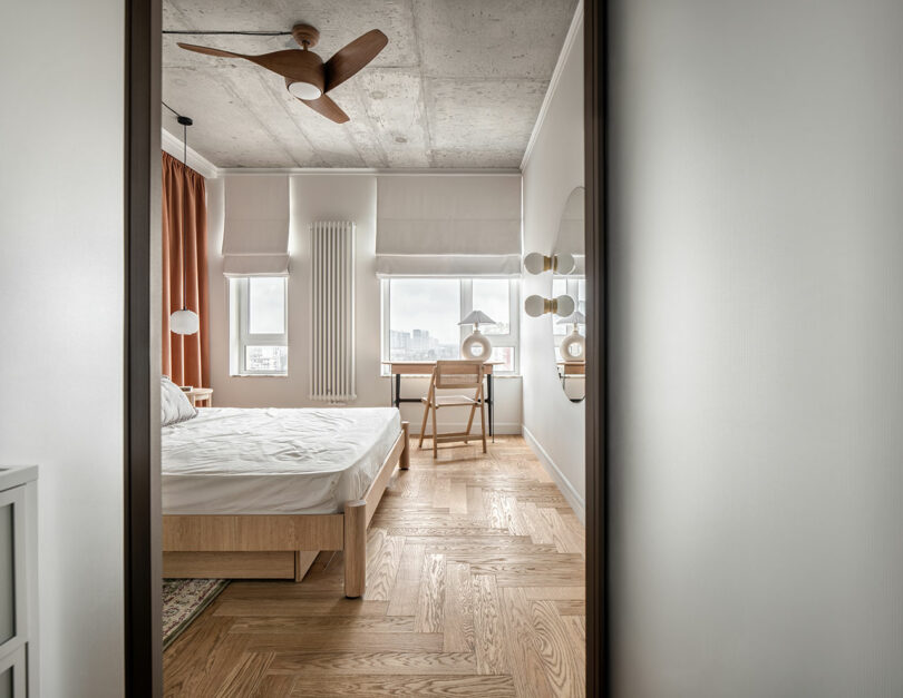 A minimalist bedroom with a wooden bed, a desk near the window, white walls, and a ceiling fan.