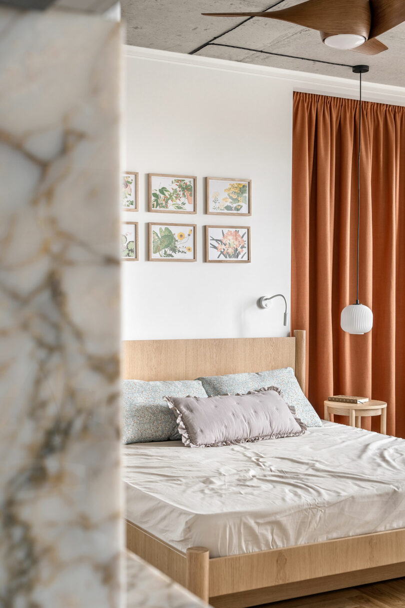 A bedroom with natural tones features a bed with neutral bedding, six botanical prints on the wall, terracotta curtains, a small nightstand with a white pendant light, and a ceiling fan.