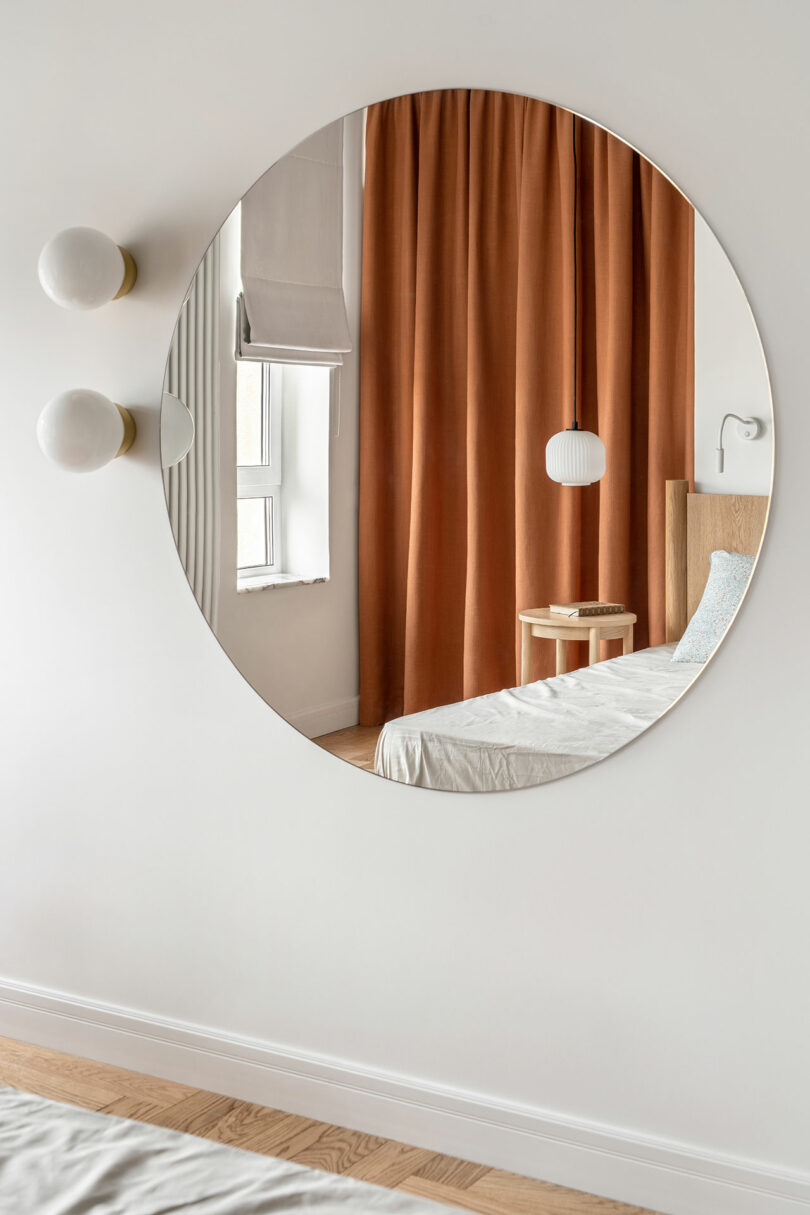 A round mirror reflecting a bedroom with orange curtains, a window with a white shade, and part of a neatly made bed with white bedding and a bedside table.