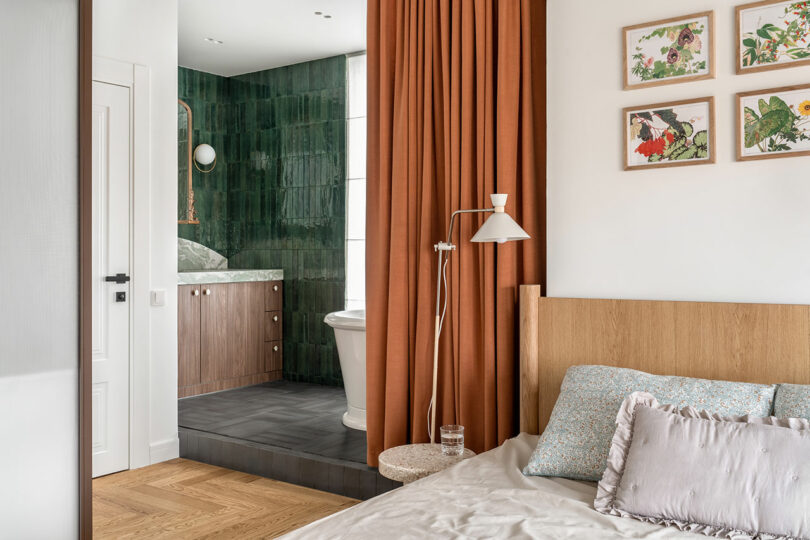 A cozy bedroom with an adjacent bathroom separated by an orange curtain. The bathroom features green tiles and a bathtub. The bedroom has a wooden bed and framed art on the wall.