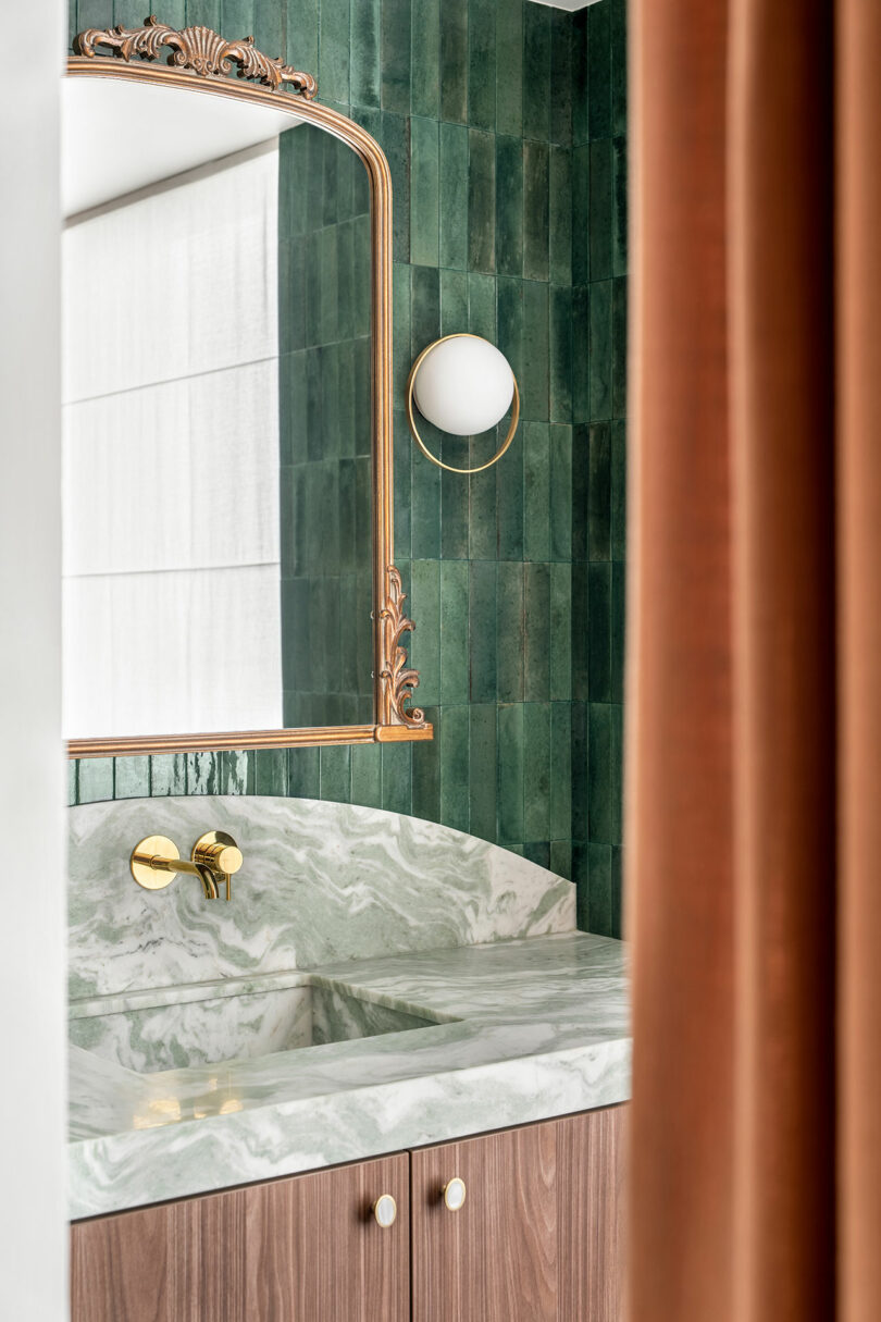 A bathroom vanity with a marble countertop, wooden cabinets, a large ornate mirror, and green tiled walls is partially obscured by a rust-colored curtain. A round light fixture is mounted on the wall.