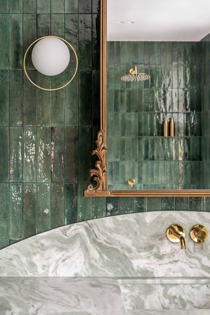 A bathroom features dark green tiled walls, matching green marble countertop, gold fixtures, and a large mirror with intricate gold edging. An elegant sconce with a round bulb is mounted on the wall.