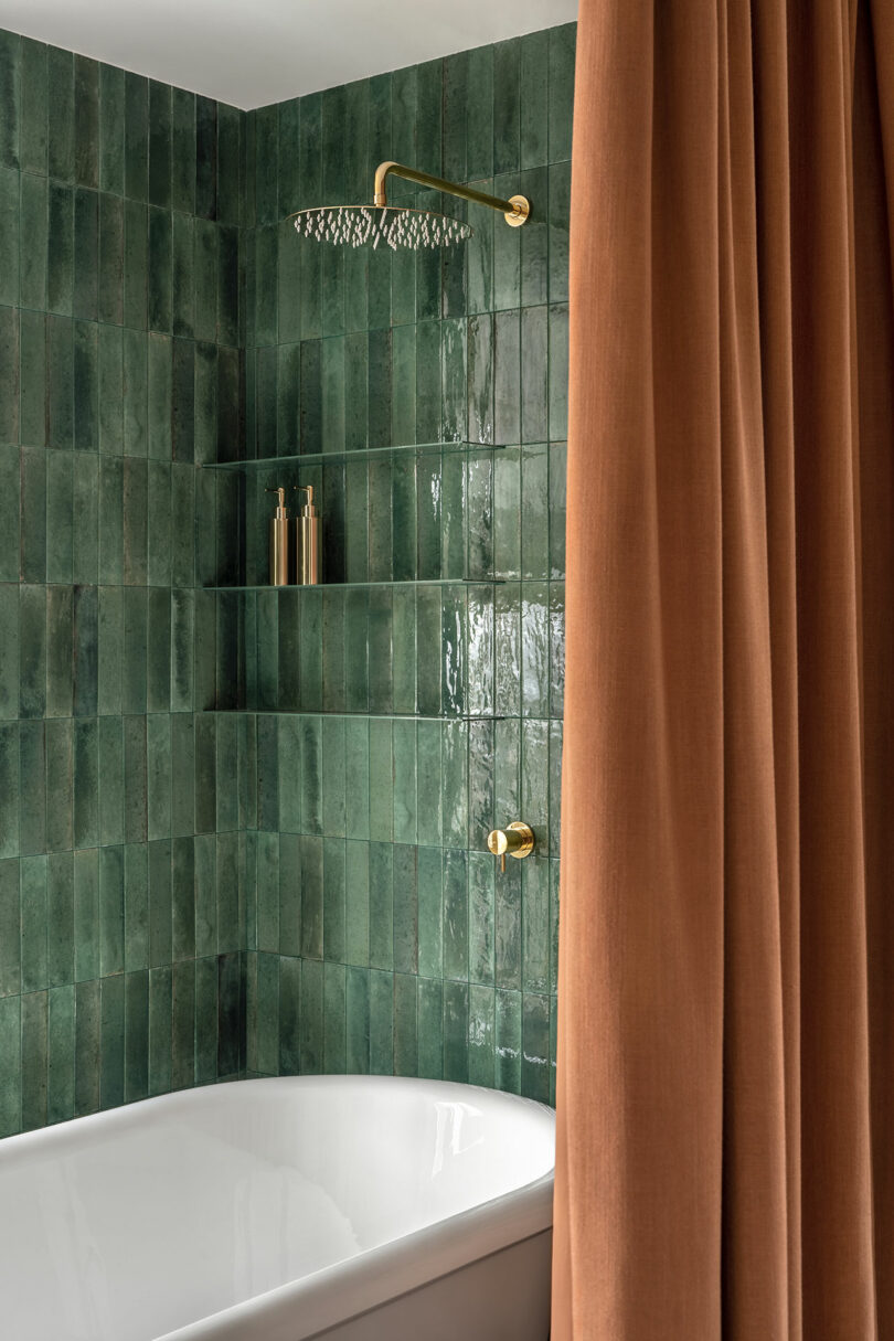 A bathroom with green tiled walls, a gold showerhead, and gold fixtures. An orange-brown shower curtain and two bottles are placed on the built-in shelf.