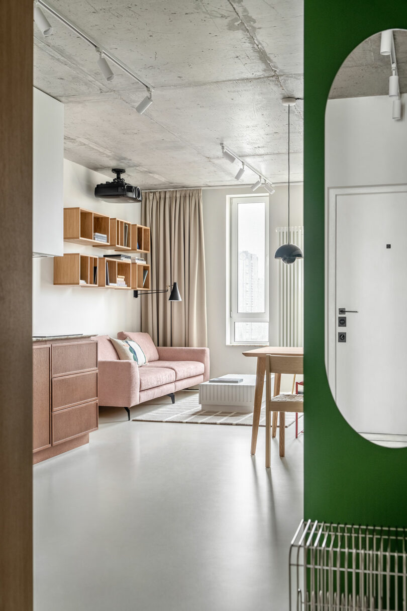 Modern living room with a pink sofa, wooden shelves, and a green archway mirror. A dining table with chairs is in the foreground, and a large window with beige curtains is in the background.