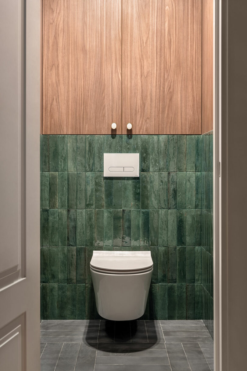 Minimalist bathroom with a green tiled wall, a white wall-mounted toilet, gray floor tiles, and wooden cabinets with two doors above the toilet.