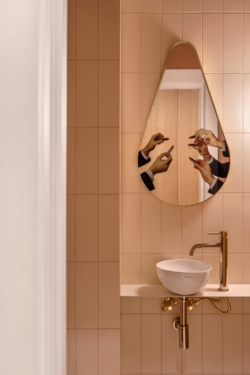 A teardrop-shaped mirror in a beige-tiled bathroom reflects a person applying lipstick. Below the mirror, a white sink and gold faucet are mounted on a thin shelf.