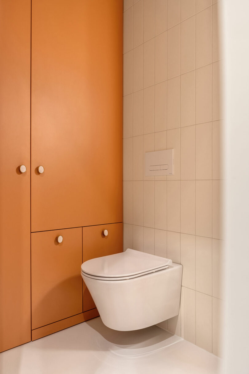 A minimalist bathroom with an orange storage cabinet and a wall-mounted white toilet against beige tiled walls.