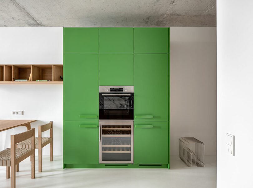 A modern kitchen with vibrant green cabinetry featuring a built-in oven. A wooden dining table with matching chairs is nearby. The space includes white walls, an exposed concrete ceiling, and minimal decor.