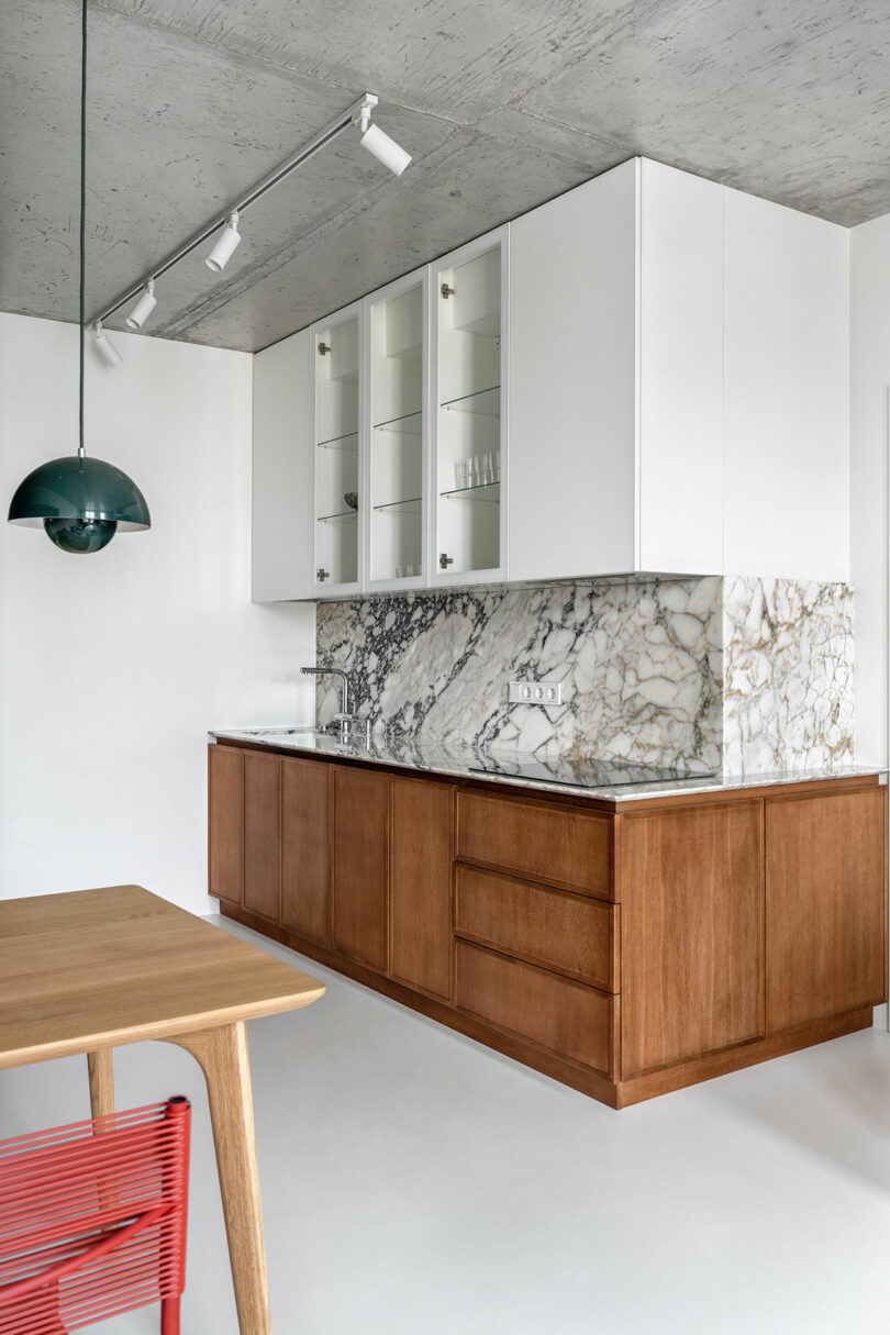 Modern kitchen with a marble backsplash and countertop, white upper cabinets, and wooden lower cabinets. A green pendant light hangs above a wooden table with a red chair.