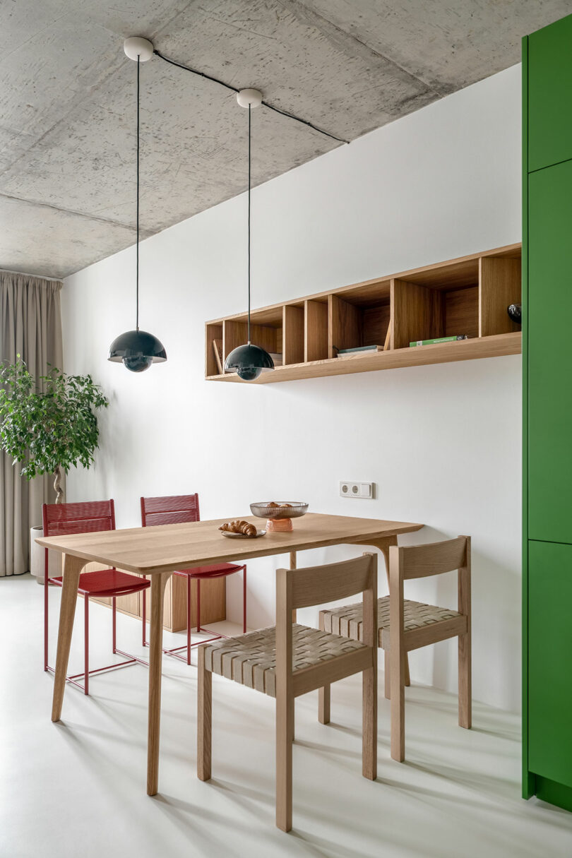 Modern dining area with a wooden table, four chairs, and pendant lights. Red chairs contrast with wooden ones. A shelf with decor items is on the wall, and a green cabinet is visible on the right.