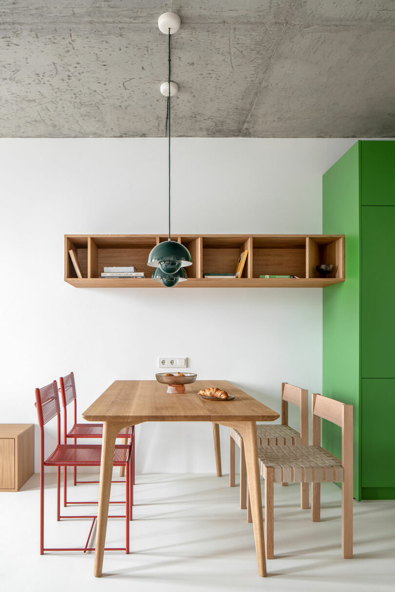 A minimalist dining area with a wooden table, red and wooden chairs, green cabinet, and hanging light. Open wooden wall shelves hold books and decor items.