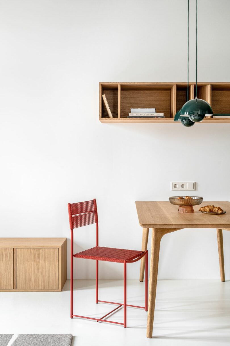 A minimalist dining area with a red chair, a wooden table, hanging shelves with books, and a green pendant light. A plate of pastries is on the table, and a wooden chest is placed against the wall.