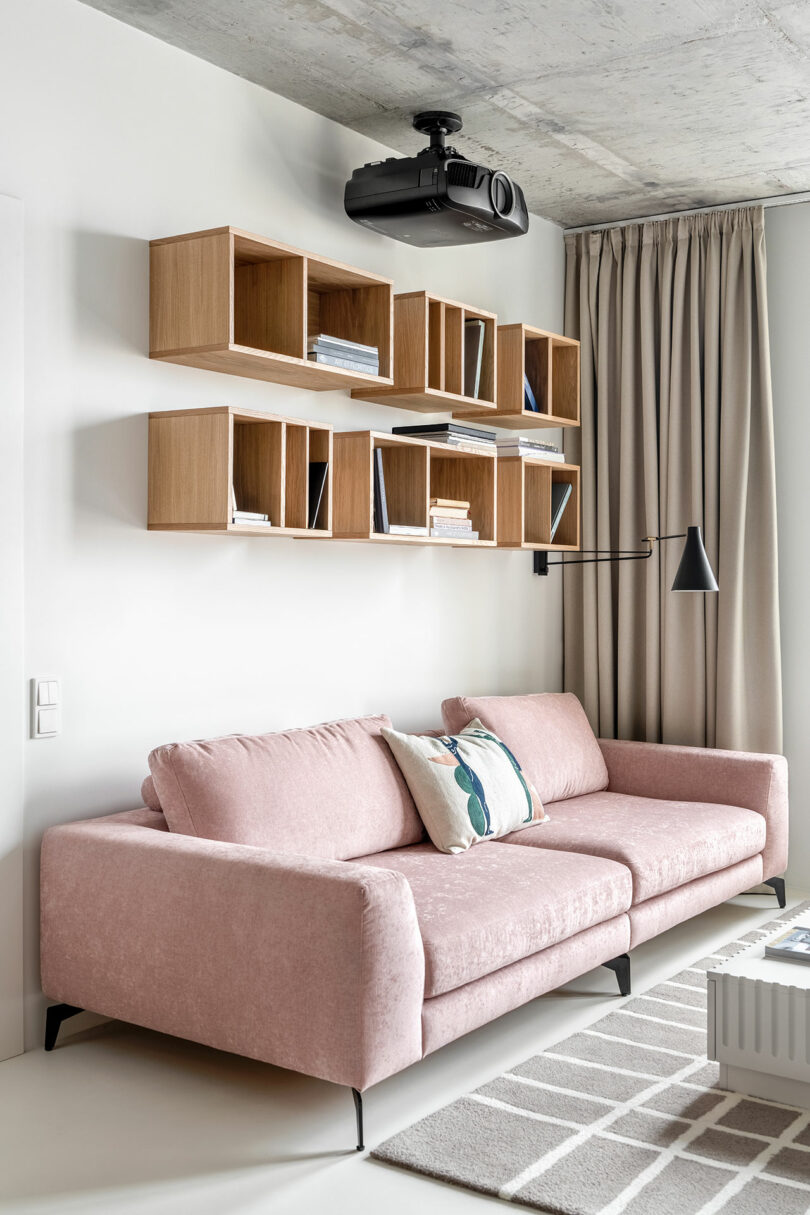 Modern living room with pink sofa, wall-mounted wooden shelves holding books, a ceiling projector, black wall lamp, and beige curtains. Geometric patterned rug on the floor.