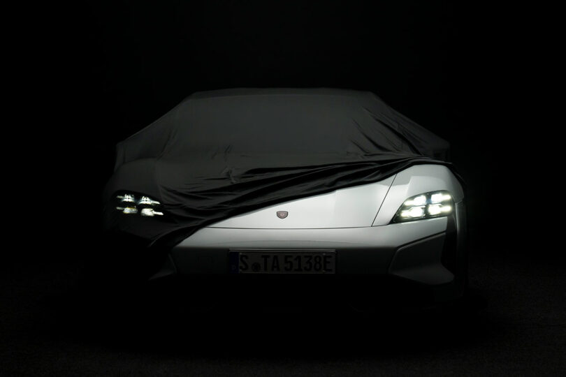 A silver car partially covered by a black cloth, showcasing its Turbonite power, is seen with its headlights on in a dark environment. Its license plate reads "S TA 5138E".