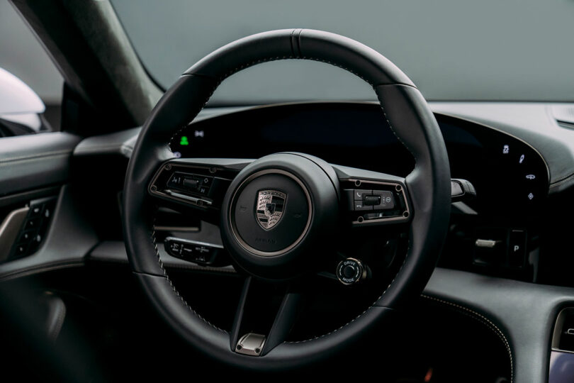 A close-up view of a black car steering wheel with the Porsche logo prominently displayed in the center. The dashboard and various control buttons are visible in the background, featuring Turbonite accents for a sleek finish.