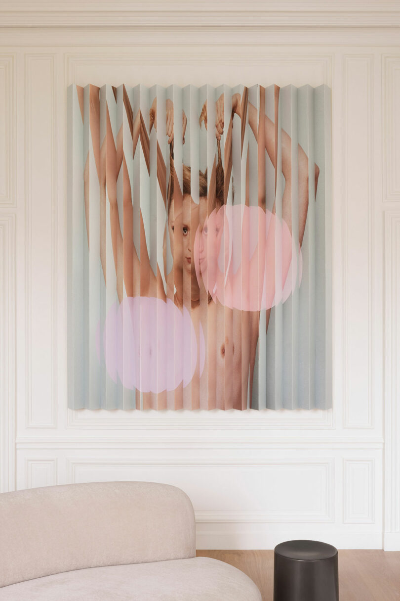 A wall installation featuring a segmented image of a person with an exposed torso, arms raised, and pink circular graphics overlaying the face. A beige curved sofa and black side table are in the foreground.