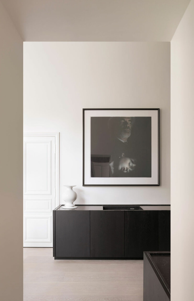 A minimalist room with light-colored walls features a black sideboard, a white door, a white vase, and a framed monochrome photograph.