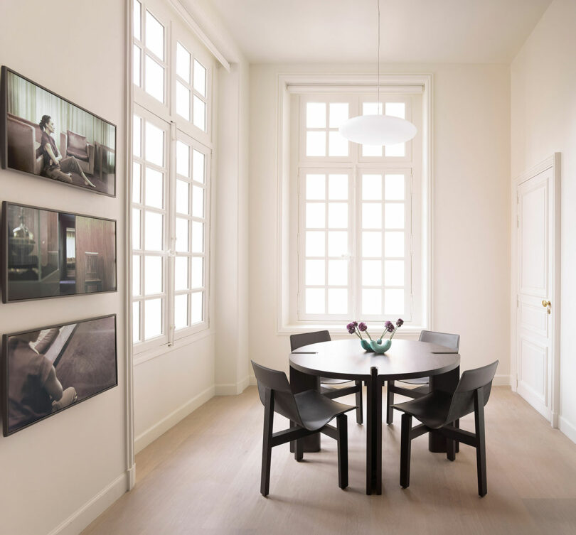 A minimalist dining room with large windows, round table, four black chairs, a vase with flowers, and three framed pictures on the wall.