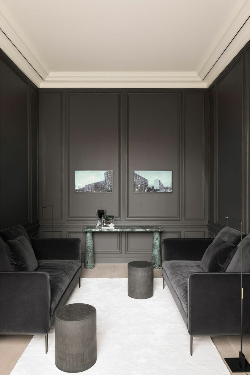 A modern living room with dark walls, two black sofas, a white carpet, and round coffee tables. A marble console table with decorative items is placed beneath two framed pictures on the wall.