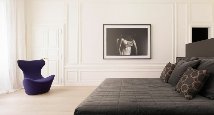 A minimalist bedroom features a black bed with patterned pillows, a purple chair in the corner, and a large black-and-white photo on the wall.