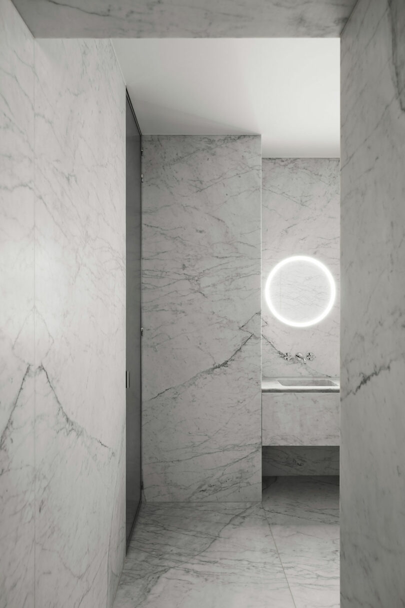 Marble bathroom with minimal design featuring a circular backlit mirror above a sink and sleek hidden storage cabinets.