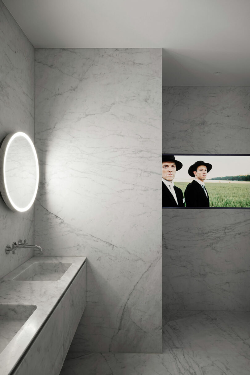 A modern bathroom with marble walls and counter, a round backlit mirror, and a recessed television screen showing two people in black attire.