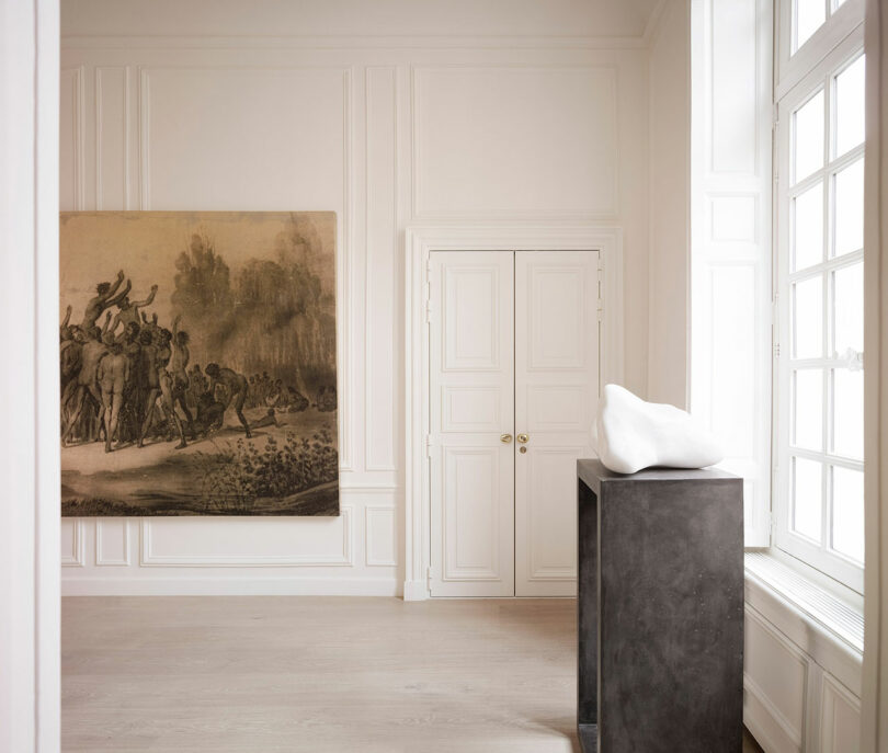 A minimalist room with white walls features a large textured painting, double doors, and a black pedestal displaying a white abstract sculpture. Natural light enters through a large window on the right.