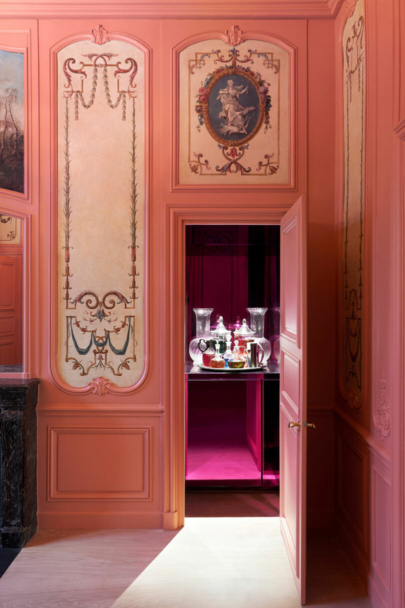 A partially open door in a room with ornate pink walls reveals a display of glass decanters and bottles on a lit-up platform in the adjacent room.