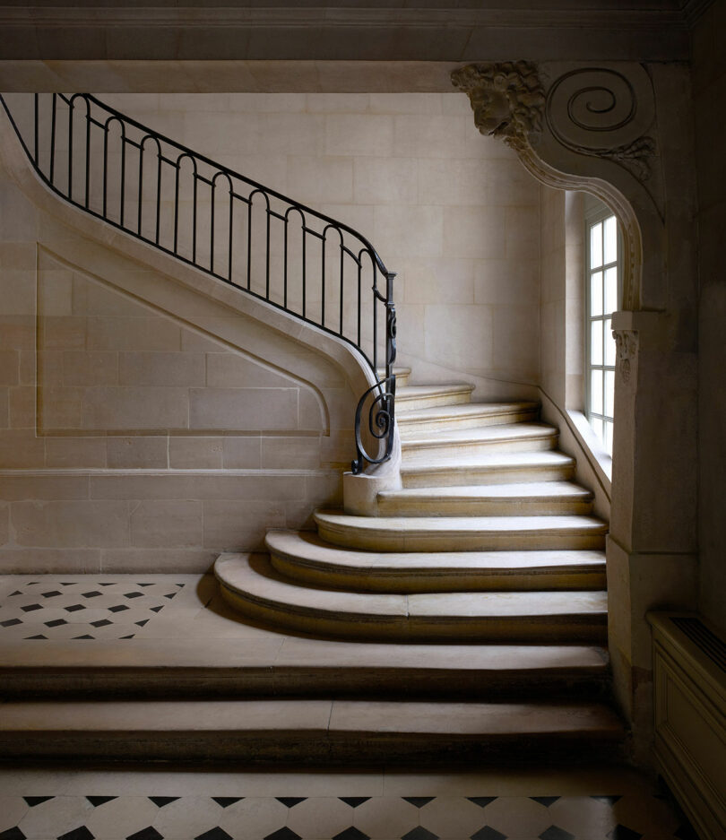 A stone staircase with a black wrought iron railing curves upwards beside a large window. The floor is tiled with a black and white checkered pattern.