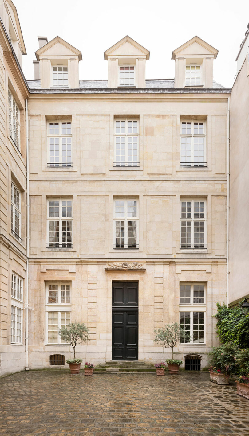 A four-story beige stone building features multiple windows with white frames and a central black door, surrounded by potted plants on a cobblestone courtyard.