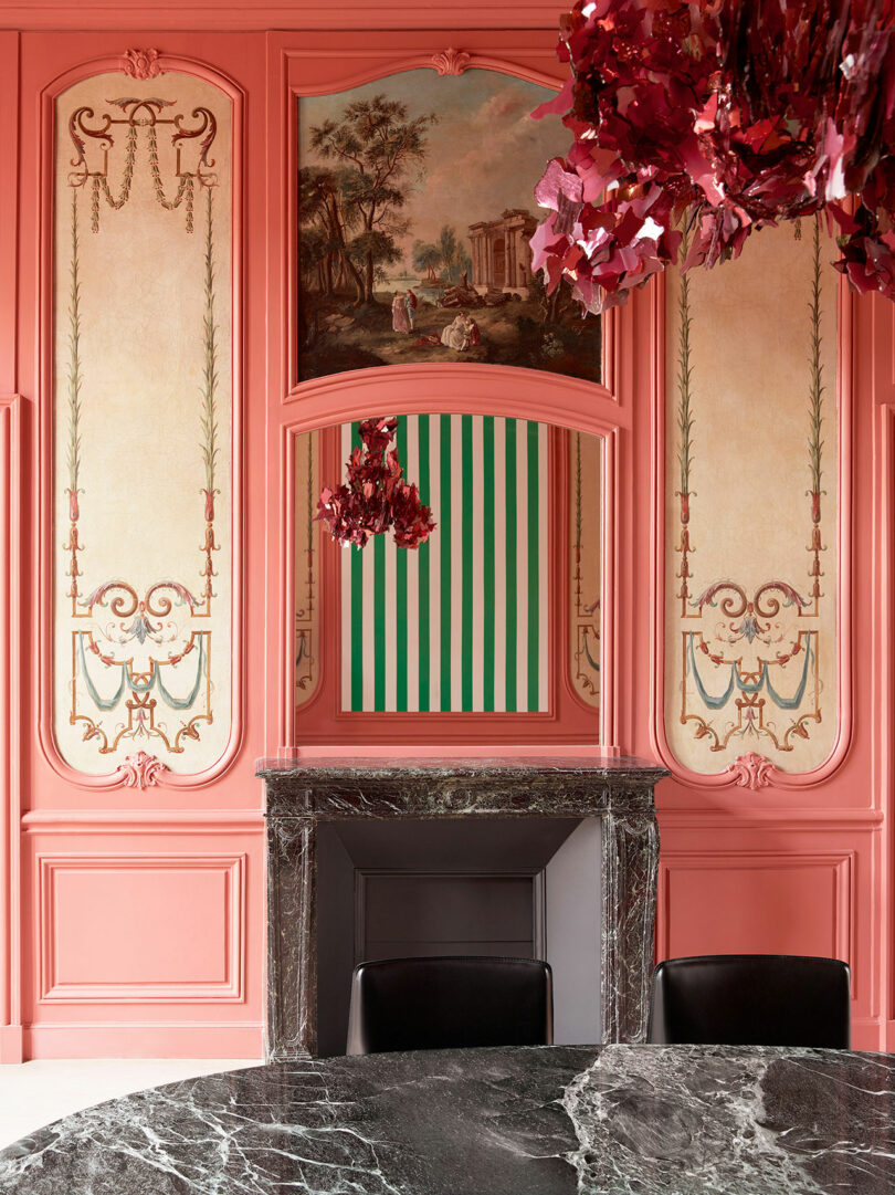 Ornate room with pink paneling, intricate decorative designs, a green and white striped wall section, and a black marble fireplace. A reddish chandelier is visible above a black marble table.