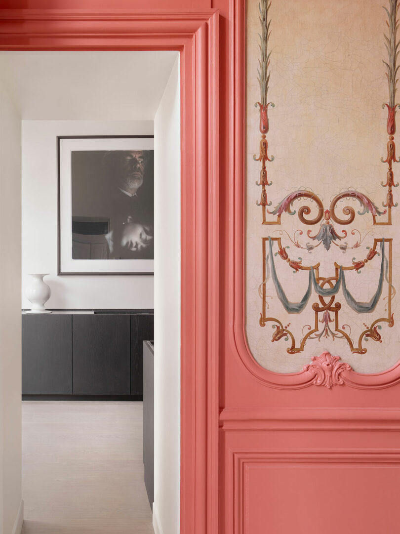 A view through a doorway with ornate pink molding, revealing a white wall with framed artwork, black cabinet, and a white vase in the background.