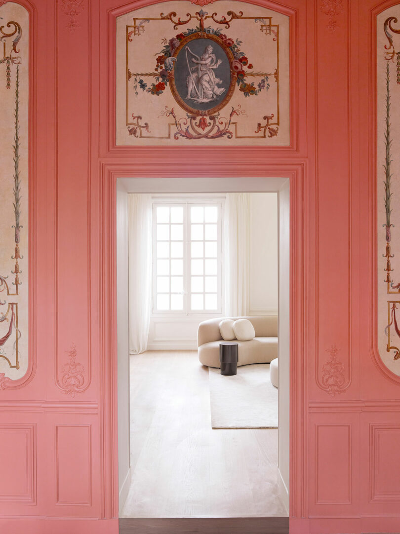 Ornate doorway in a pink wall with classical artwork leads to a minimalist room featuring a light-colored sofa, white curtains, and a small dark table. Light floods in through a large window.