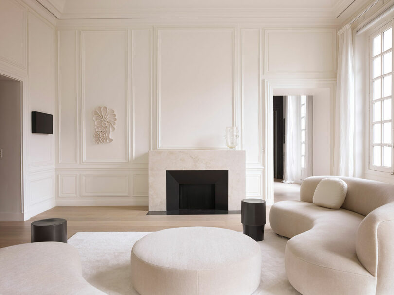 A minimalist living room features a modern sofa, round coffee tables, and a decorative fireplace with a marble facade against white walls and large windows.