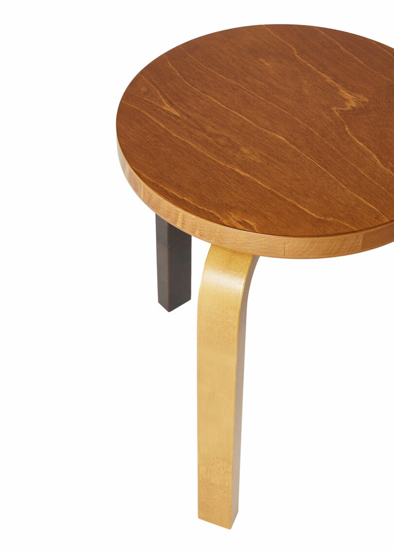 Stool 60 by Alvar Aalto with a smooth top and three curved legs, one of which is a different color.