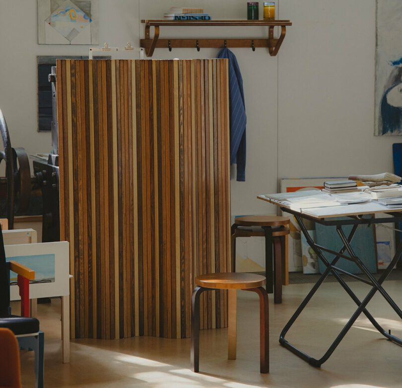 A room with a wooden foldable divider, a brown stool from Paul Smith, a desk with stacked books and papers, and shelves with various items. Artek artworks are visible on the walls and a striped cloth hangs on a hook.